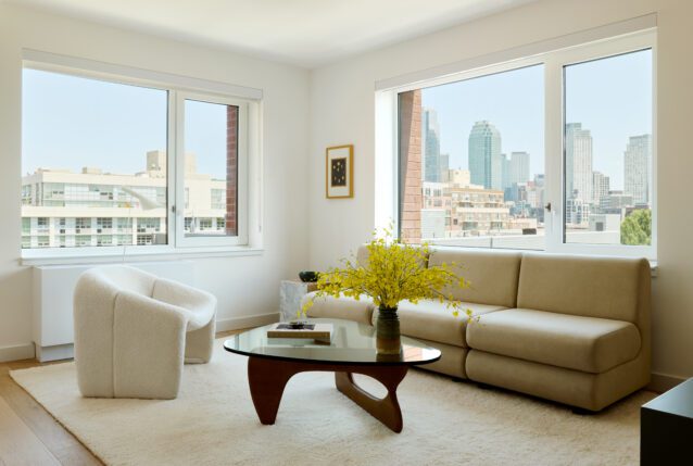 A corner living room with a tan couch, white chair, and windowed views of Long Island City